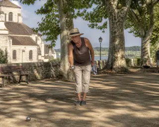 Pétanque player ready to throw on a sunlit dirt boulodrome under plane trees near a historic church in Saint-Leu-d’Esserent, with a golden Mediterranean atmosphere.